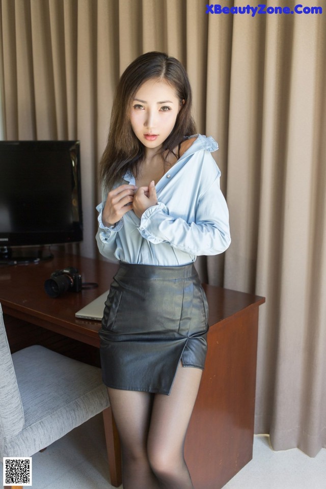 A woman in a blue shirt and black skirt posing in front of a desk.